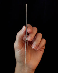A hand holds a sterling silver wand necklace pointed up against a black background. The wand is polished silver and square shaped, with two silver rings to define the top and bottom of the handle portion of the wand.
