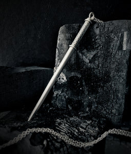 A sterling silver wand necklace lays against a black charcoal block. The wand handle has a hand-engraved floral pattern, with a silver ring at the top and bottom of the engraving.